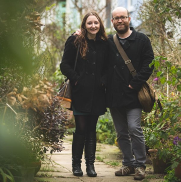 Kate is a White woman with long fair hair and Mark is a White man with a beard and glasses. They are standing in a garden